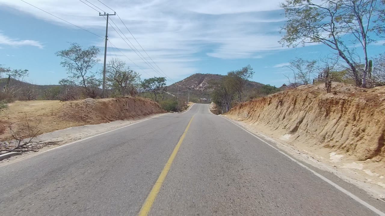 The road continues through East Cape Los Cabos with a sandy bank close on the right, sparse brush and cactus along both sides, utility poles overhead, distant hills, and no visible people.