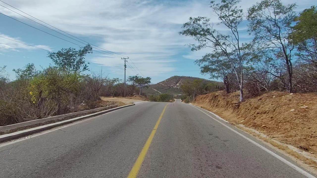 Road-level view in East Cape Los Cabos, Baja California Sur, showing a paved two-lane road, yellow center line, dry roadside brush, utility poles, distant hills, and no visible people.