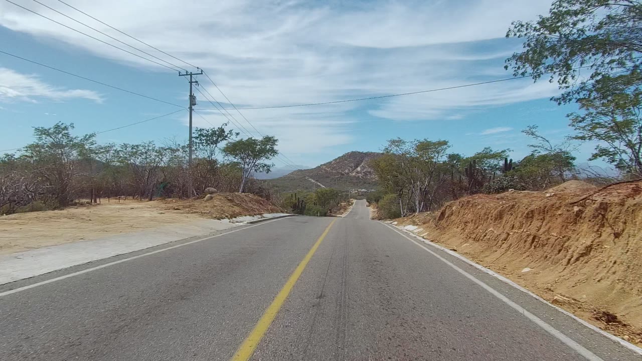 Paved road with yellow center line, dirt shoulder, trees, and low hill ahead.