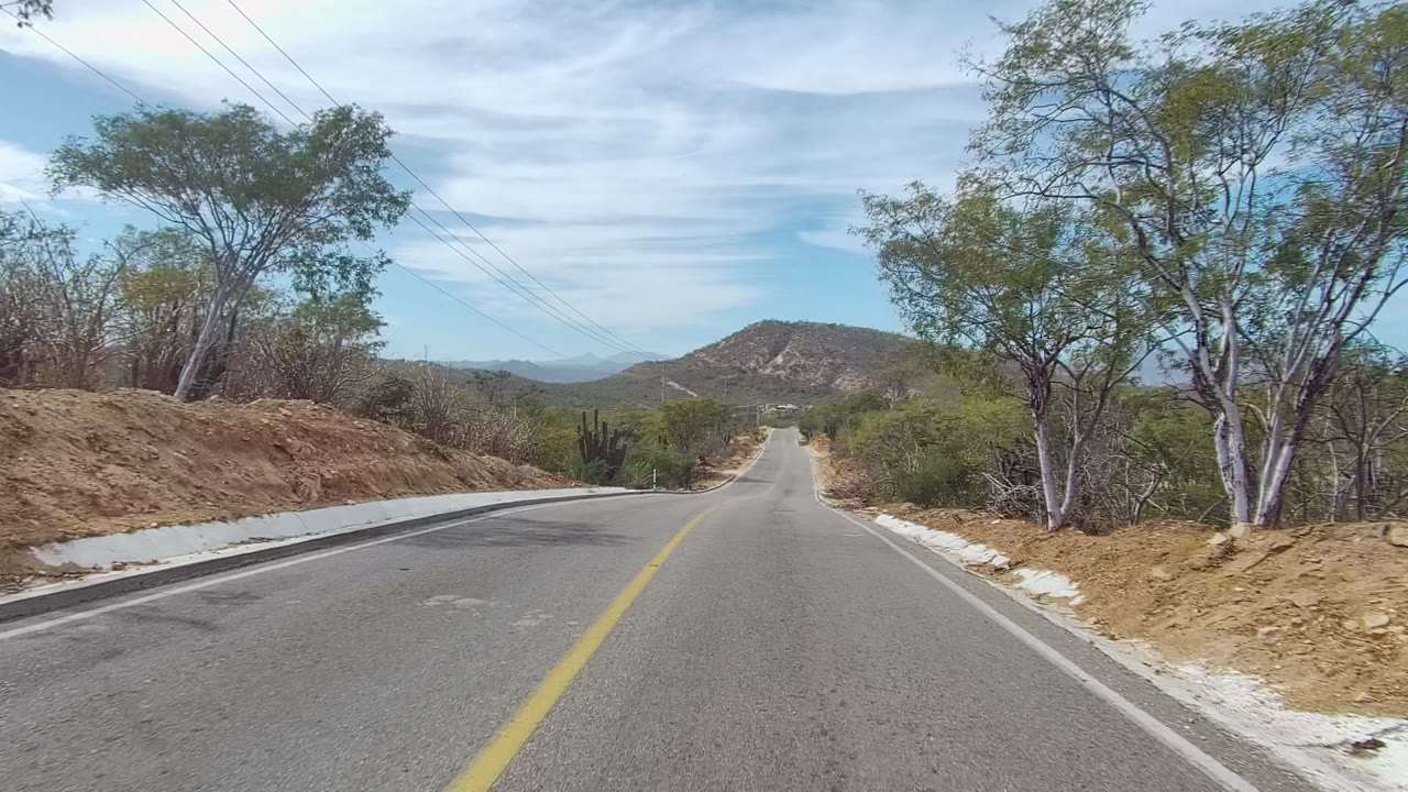Road-level East Cape Los Cabos frame showing a slight bend, raised dirt embankment, sparse roadside trees, overhead power lines, distant hills, and no visible people.