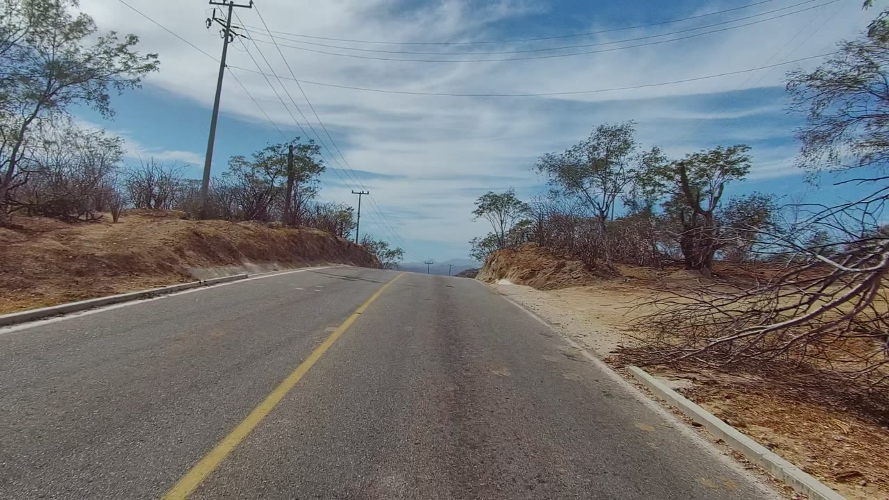 Start of the East Cape Los Cabos road clip, with a paved lane, yellow centerline, dry embankments, sparse trees, utility poles, power lines, blue cloudy sky, and no visible people.