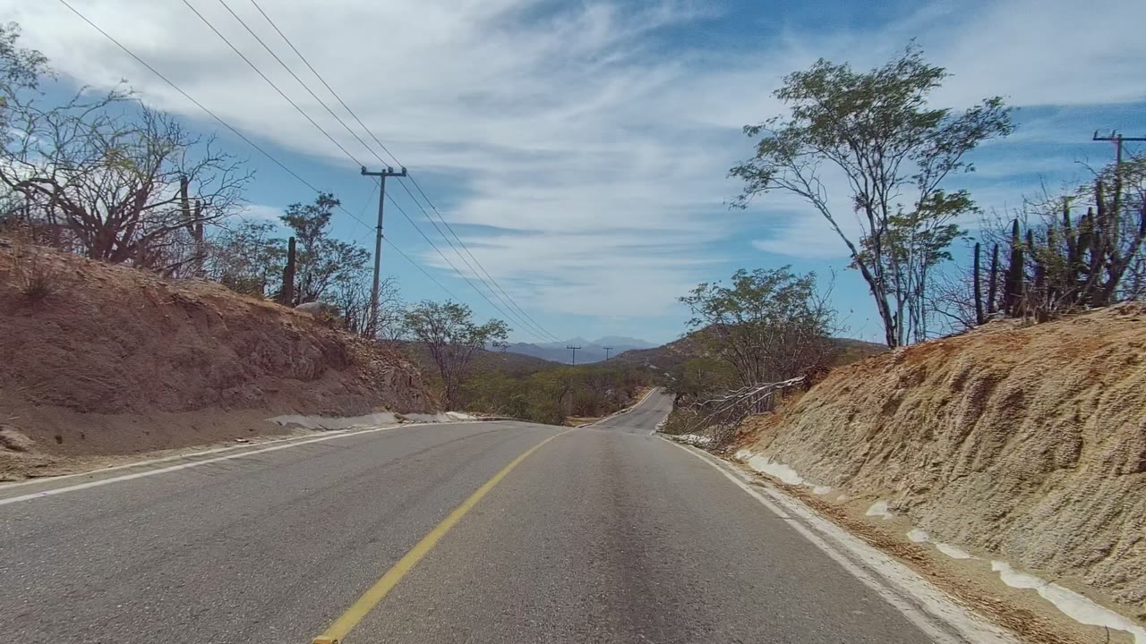 At 0:02, the East Cape Los Cabos road bends past dry tan slopes, leafless brush, sparse green trees, roadside drainage, distant hills, power lines, and no visible people.