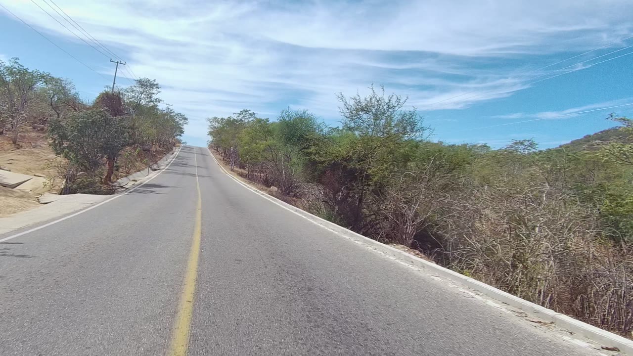 Forward view of a paved road in East Cape Los Cabos, Baja California Sur, with dry brush on both sides, utility lines at left, blue sky with thin clouds, and no visible people.