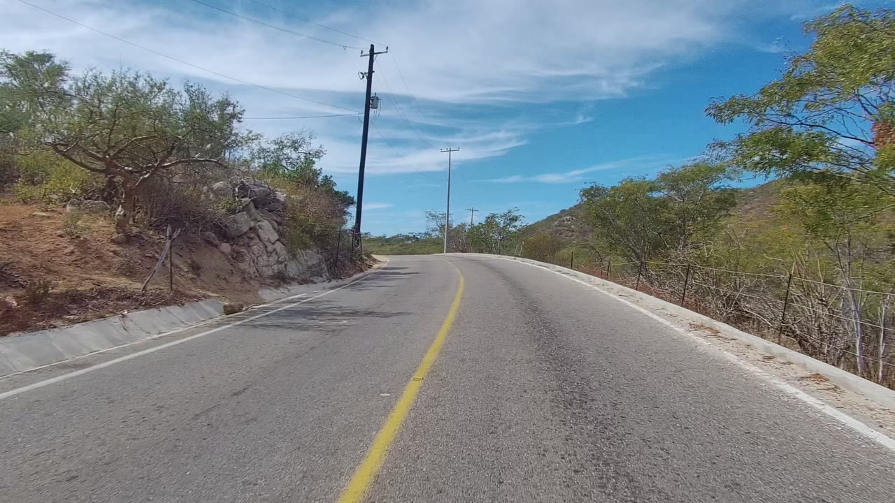 East Cape Los Cabos road view with a yellow center line, scrub trees, rocky roadside cuts, utility poles, blue sky, and no visible people.