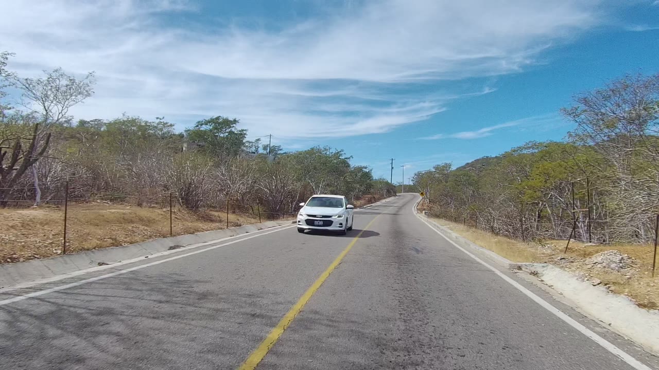 A rural road in East Cape Los Cabos, Baja California Sur, with a white car approaching on the left side of the frame, dry roadside trees, a yellow center line, and no visible people.