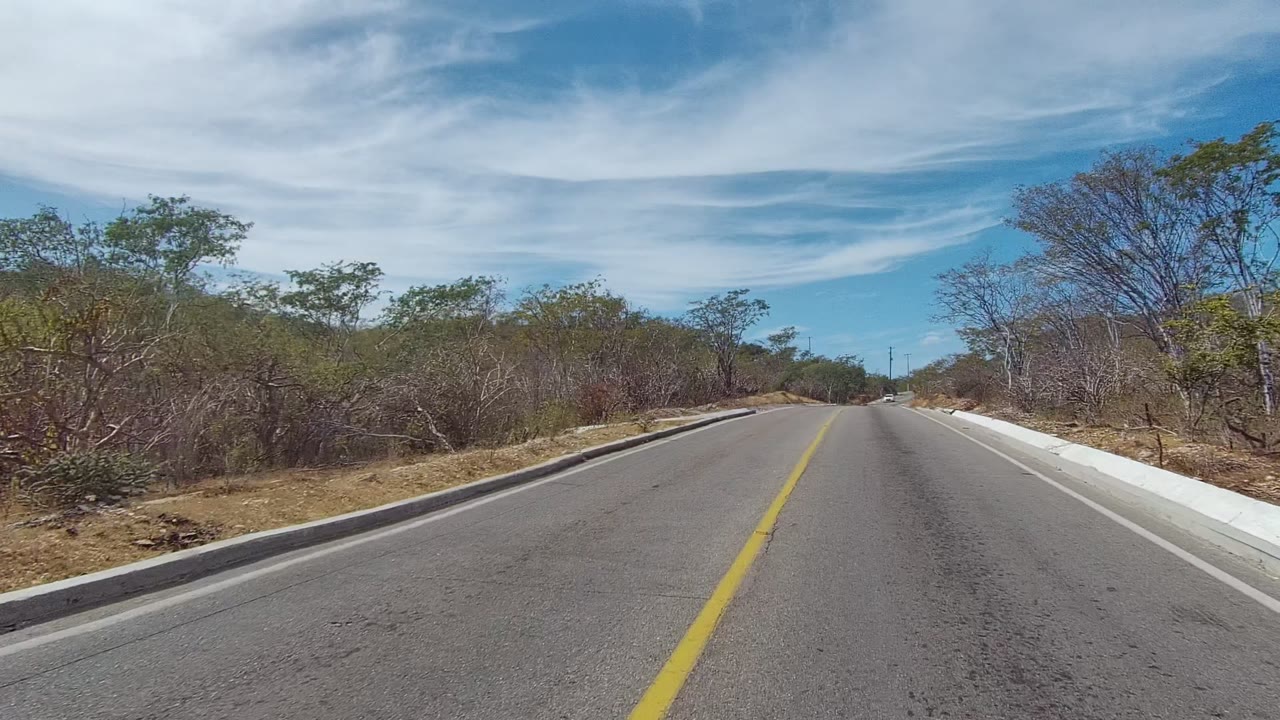 East Cape Los Cabos road scene with the lane stretching ahead, dry scrub along both sides, a bright white curb on the right, wispy cloud bands, and no visible people.