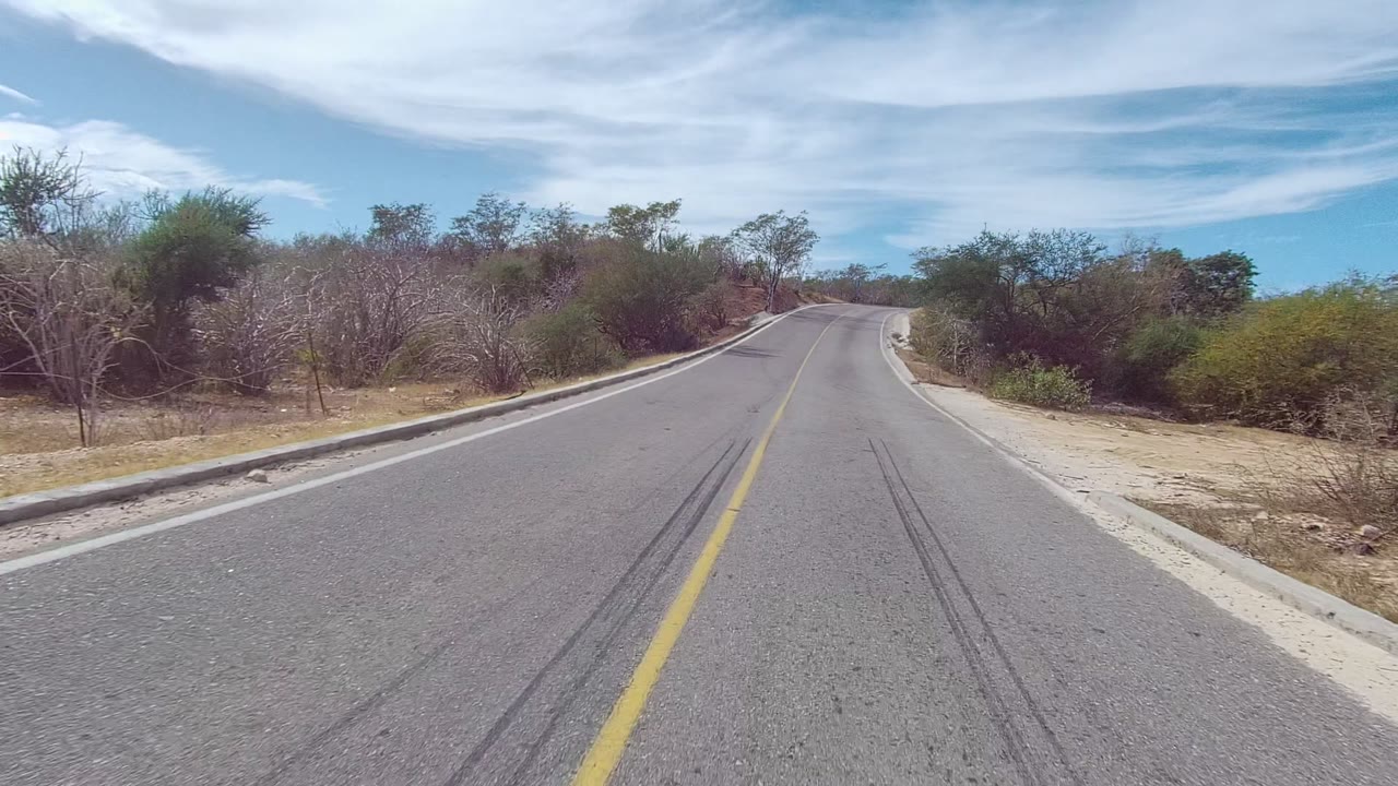A paved East Cape Los Cabos road with a yellow center line runs ahead through dry brush and small trees under a partly cloudy blue sky, with no visible people.