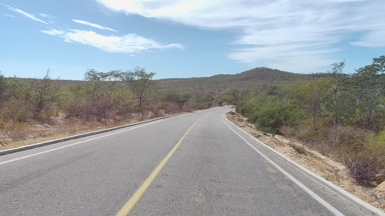 A paved road in East Cape Los Cabos curves gently right through dry shrubs and low hills under streaked white clouds, with no visible people.