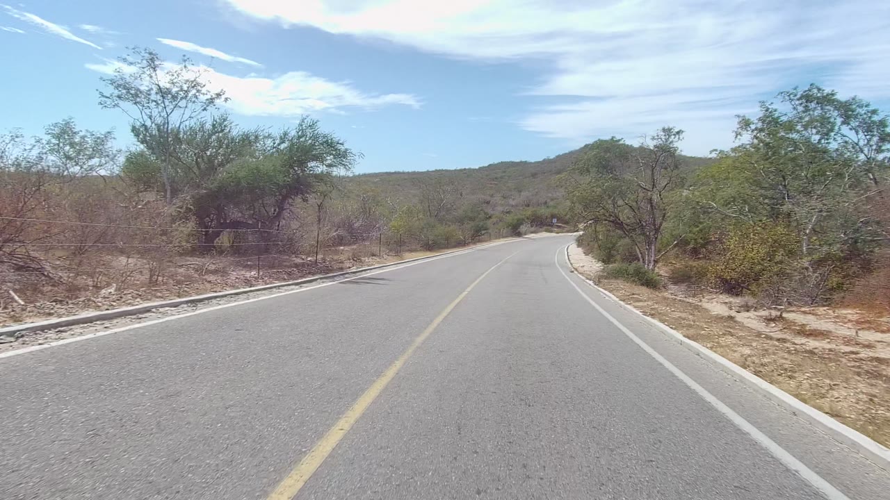 The East Cape Los Cabos road bends right with dense greenish shrubs on the shoulder, low hills ahead, and bright clouded sky, with no visible people.
