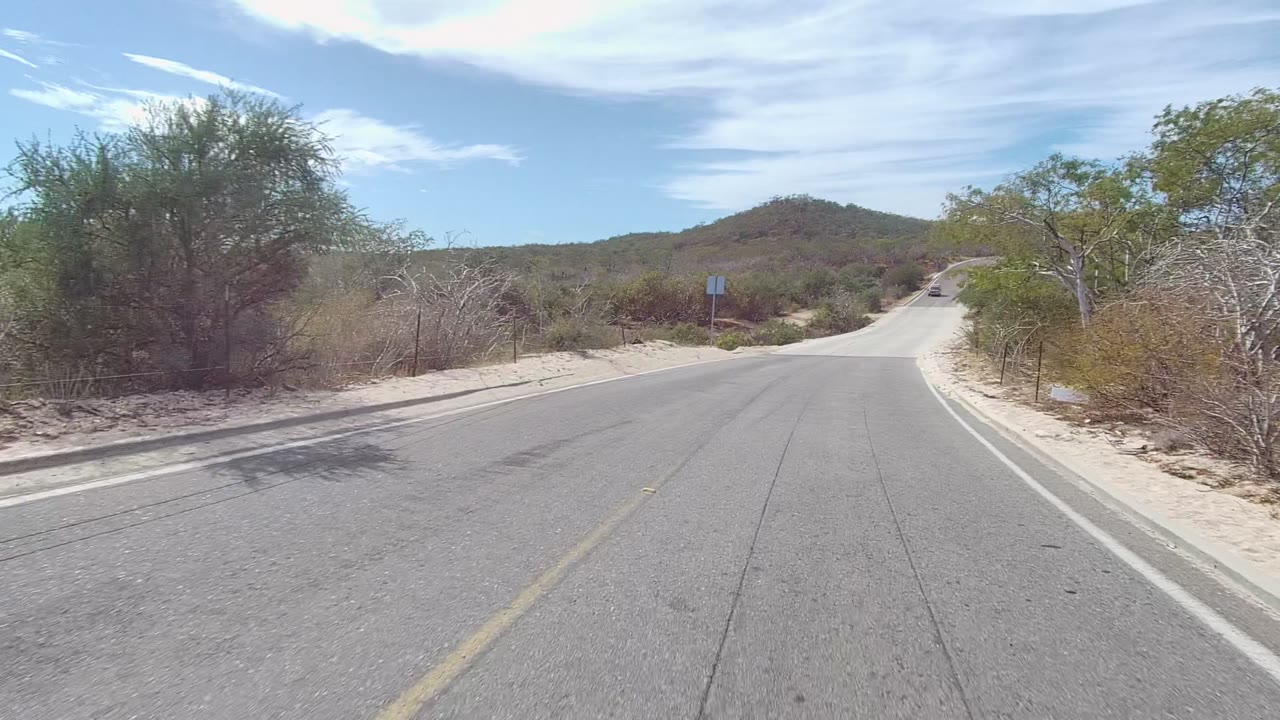 East Cape Los Cabos paved desert road bordered by pale sand, sparse green shrubs, low hills, and streaked clouds, with no visible people.