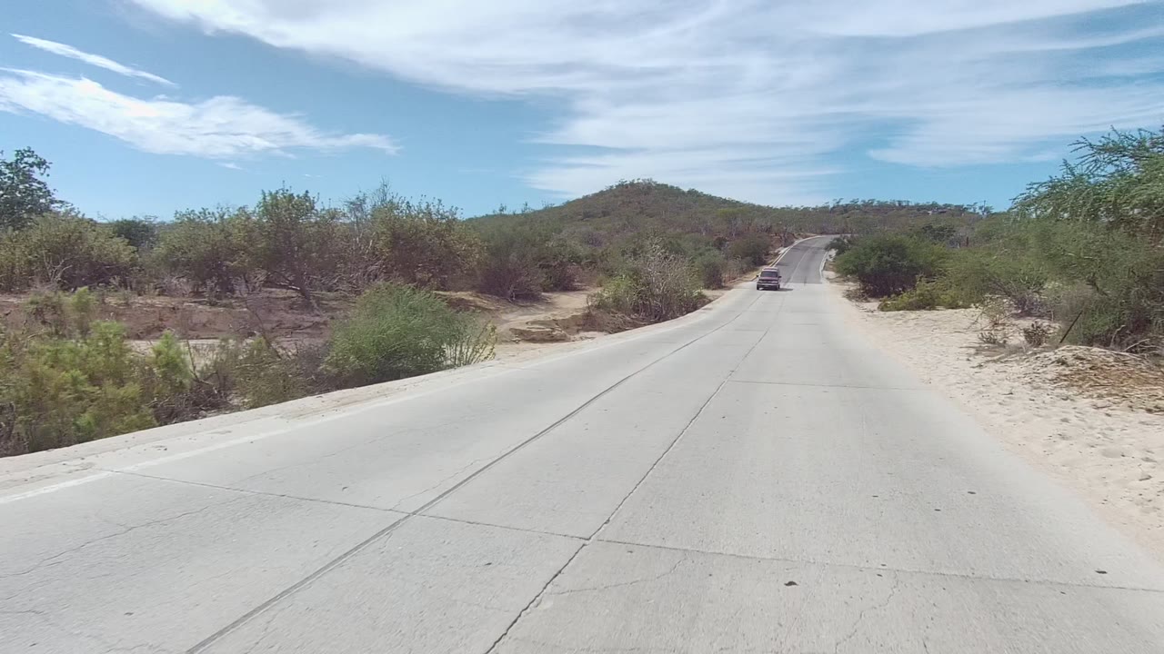 East Cape Los Cabos desert road with a vehicle ahead in the lane, sandy shoulders, scrub vegetation, low hills, and no visible people.