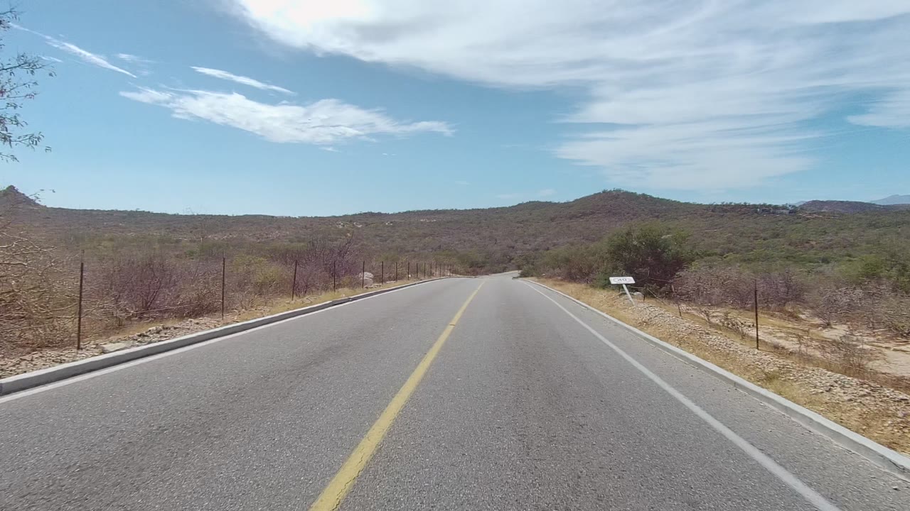 Straight two-lane desert road with low hills and partly cloudy sky