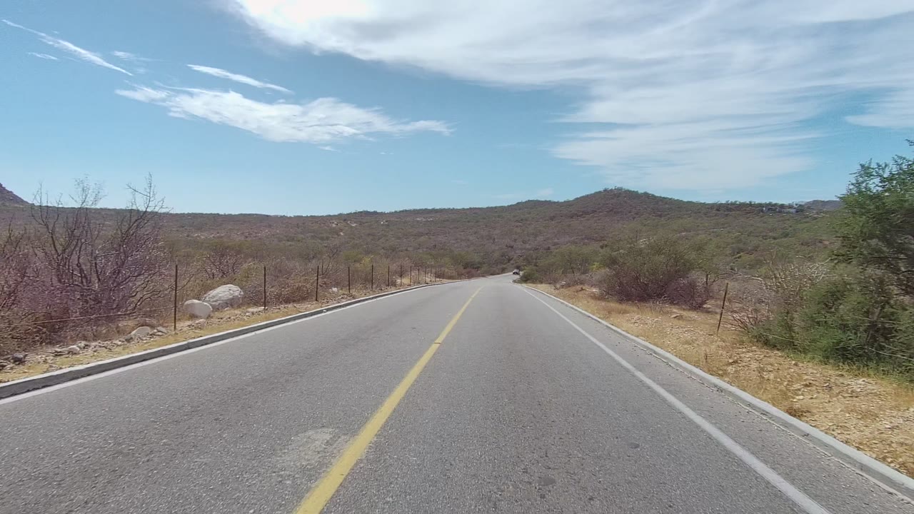 The Los Cabos highway remains centered, bordered by dry vegetation and fencing, with low hills ahead and no visible people in frame.
