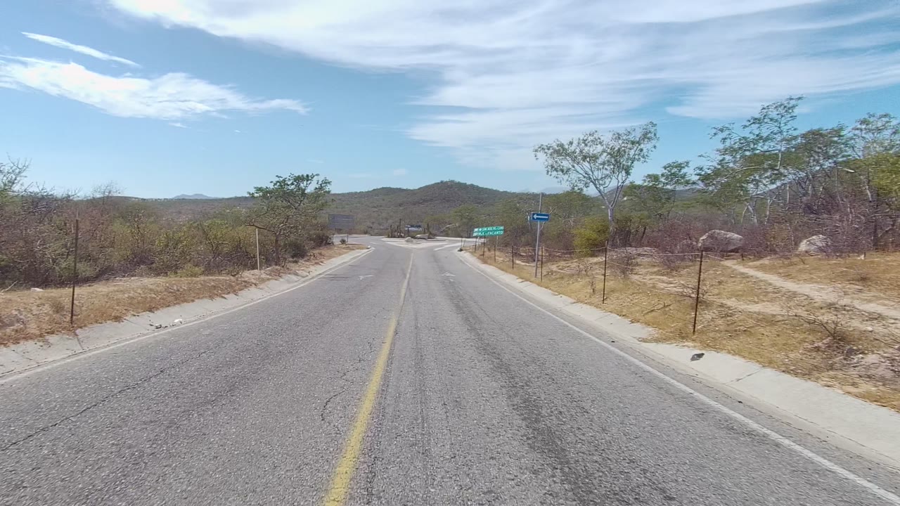 A paved road in East Cape Los Cabos stretches ahead through dry scrub and low hills, with a green roadside sign in the distance and no visible people.