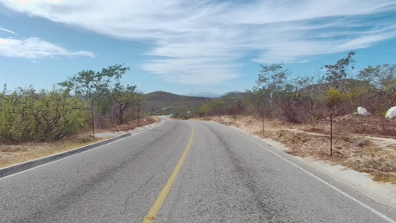 Start of a forward road view in East Cape Los Cabos, with a yellow center line, dry roadside brush, low hills ahead, and no visible people.