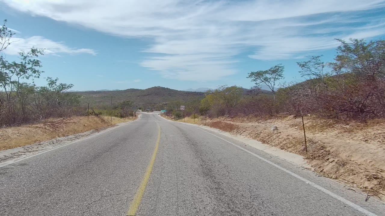 The road in East Cape Los Cabos continues through sparse brush, with a visible sign near the right shoulder, low hills ahead, and no visible people.