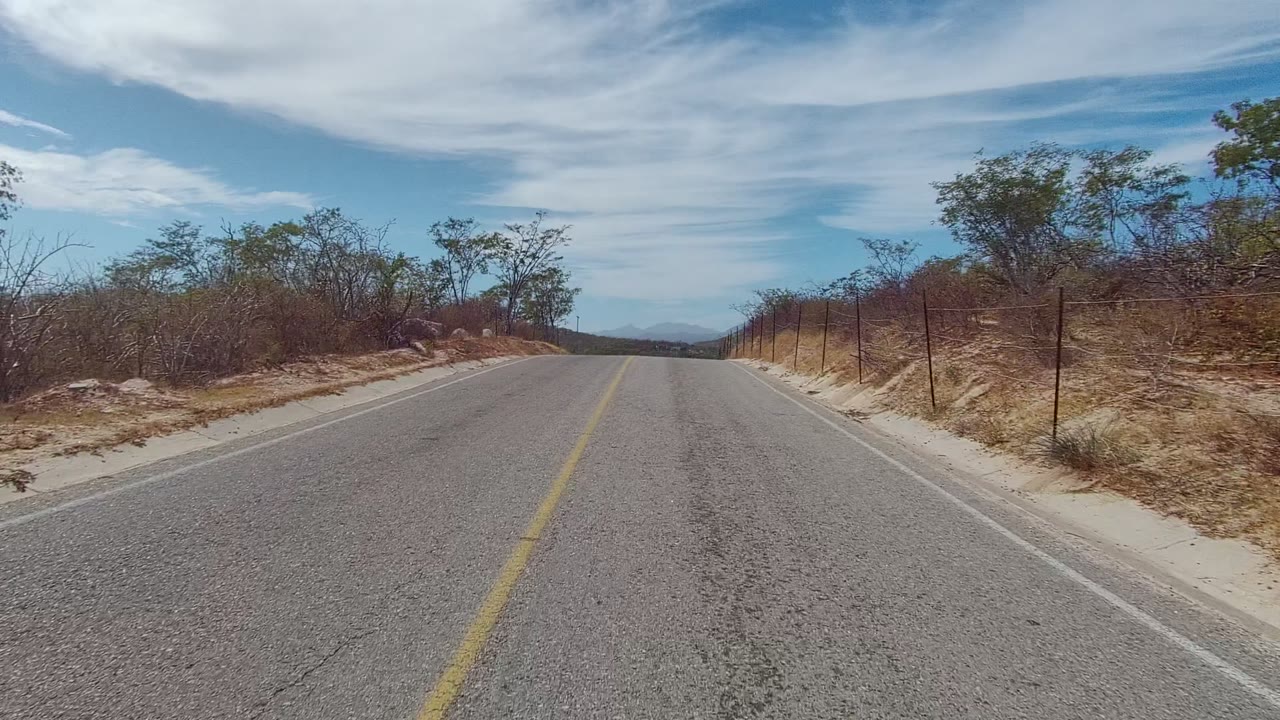 Start of the East Cape Los Cabos road clip showing a paved road, yellow center line, sandy shoulder, wire fence, dry brush, distant hills, cloudy blue sky, and no visible people.