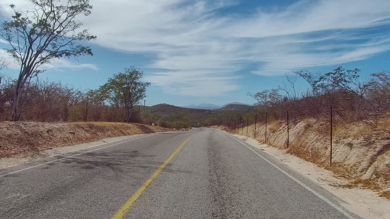 East Cape Los Cabos road scene at 0:02 with asphalt filling the foreground, a yellow center line, sandy banks, sparse trees, distant rolling hills, high clouds, and no visible people.