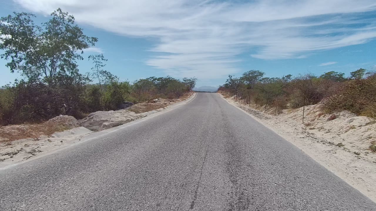 East Cape Los Cabos roadway continues through sparse scrub with pale sandy shoulders, a fence line, distant hills, and broad blue sky, with no visible people.