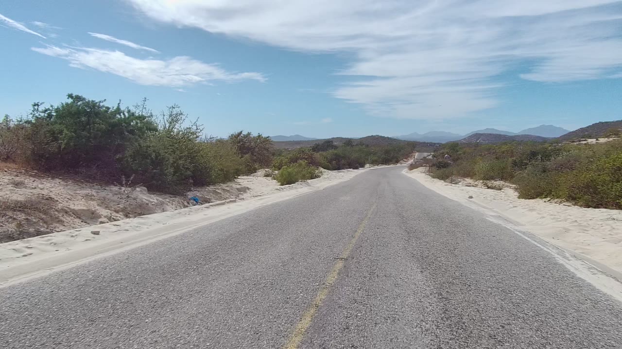 Opening frame of a paved road in East Cape Los Cabos, Baja California Sur, with sandy shoulders, low shrubs, distant hills, streaked clouds, and no visible people.