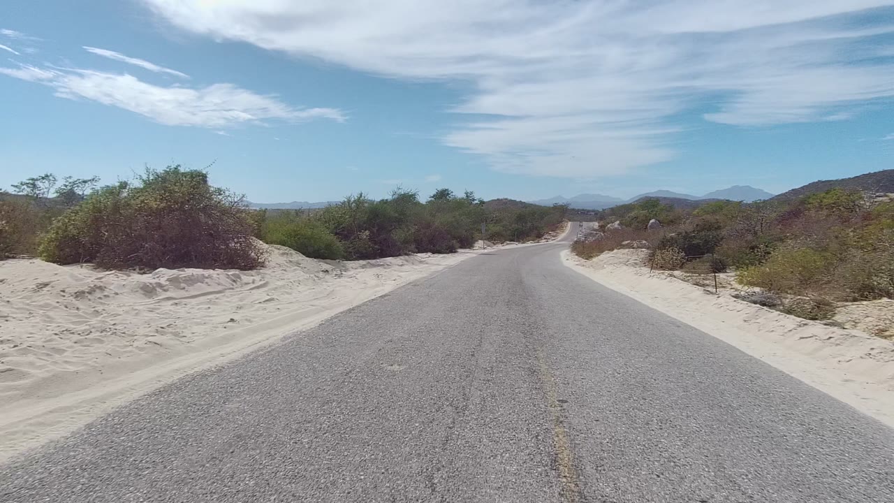 The East Cape Los Cabos road continues with a slight curve ahead, sandy ground tight to both edges, green shrubs, distant ridges, high clouds, and no visible people.