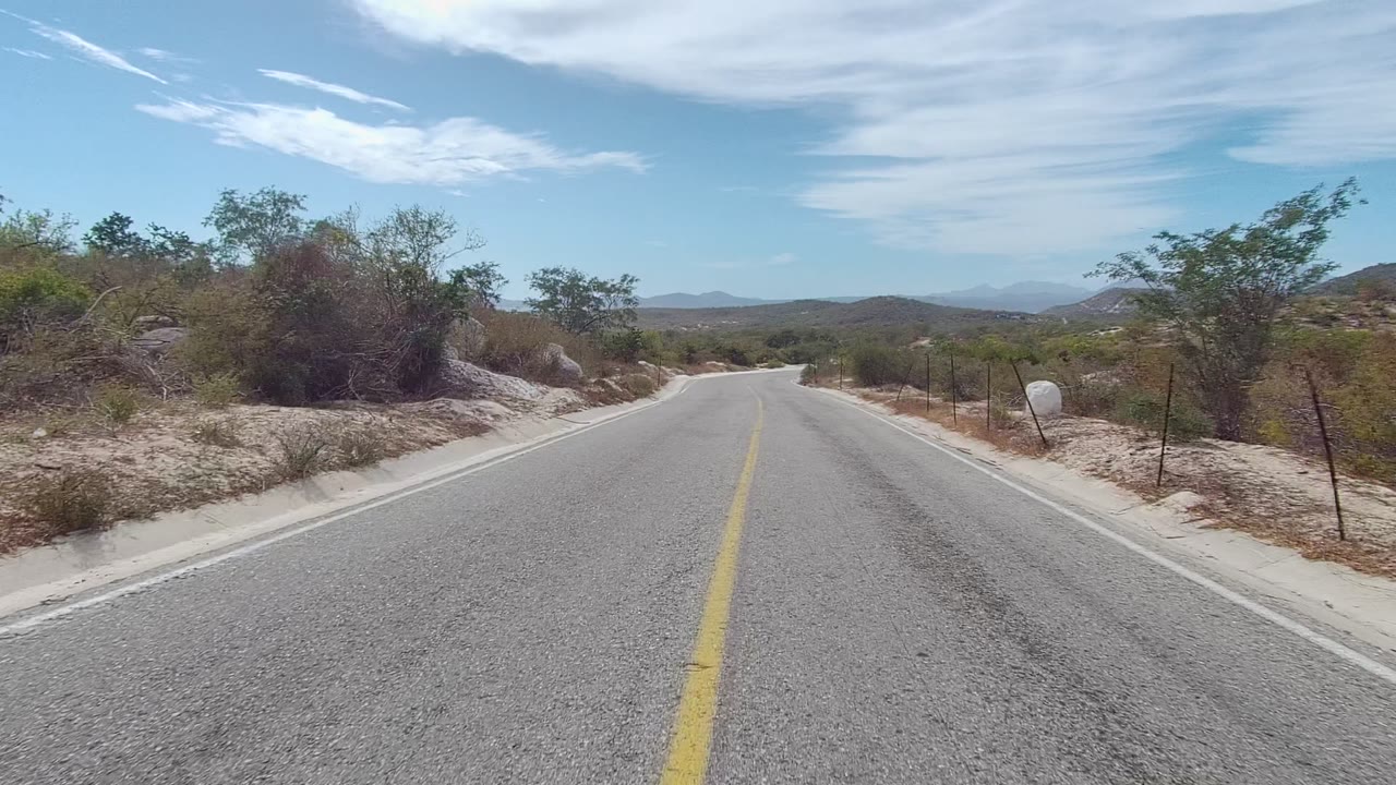 A paved road in East Cape Los Cabos, Baja California Sur curves through dry brush and rocks under a blue sky, with no visible people.