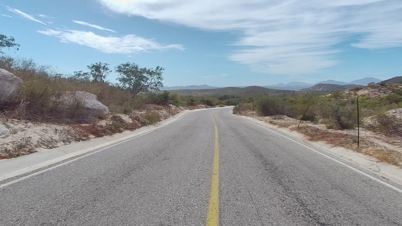 Empty paved road with roadside shrubs and distant hills.