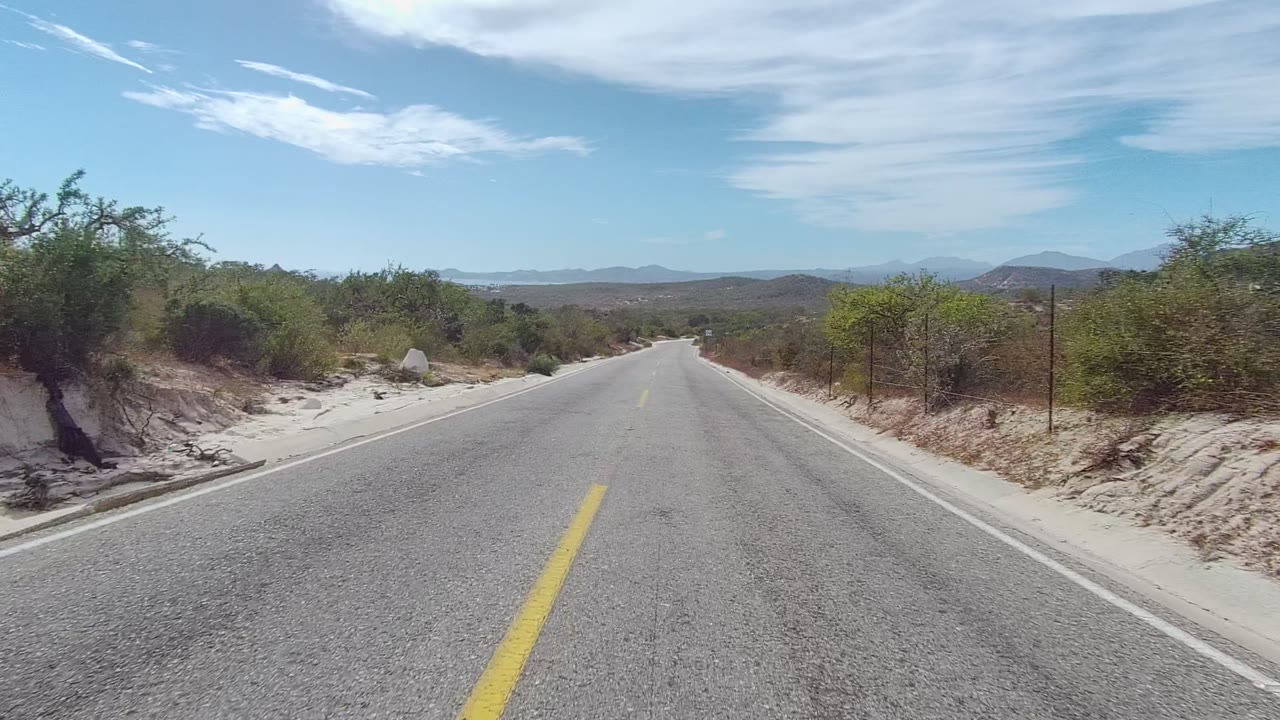 Opening frame of a straight paved road in East Cape Los Cabos, Baja California Sur, with a yellow center line, dry roadside scrub, distant hills, blue sky, and no visible people.