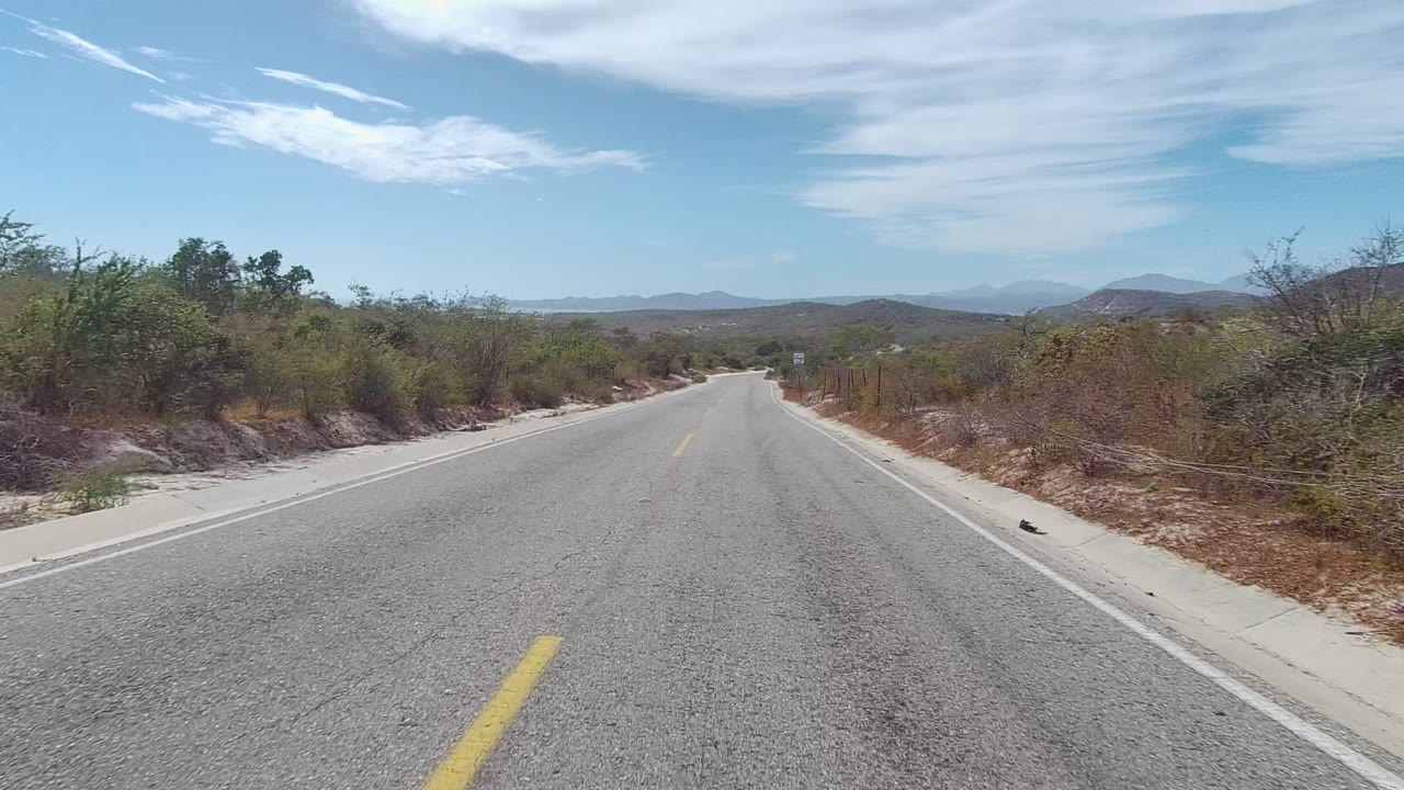 Third frame looking down the paved road in East Cape Los Cabos, with scrub on both sides, low mountains on the horizon, wispy white clouds, and no visible people.