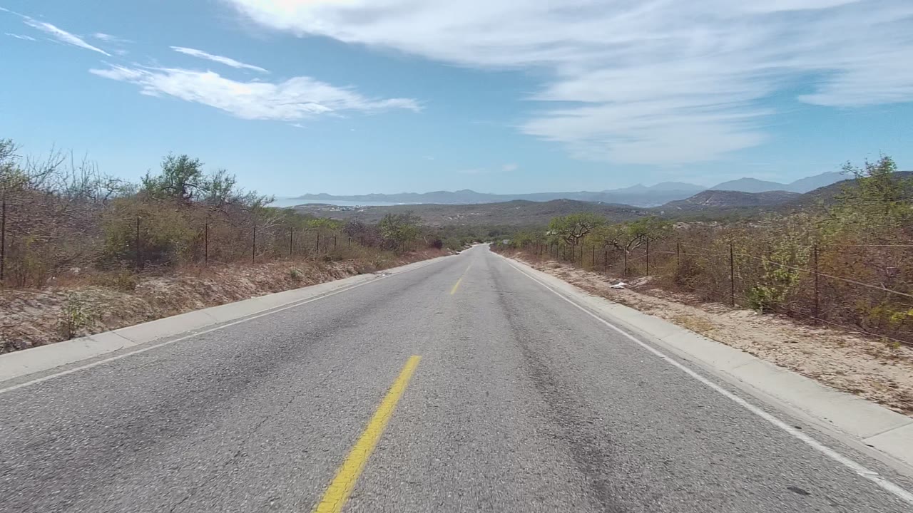 East Cape Los Cabos two-lane asphalt road stretches forward between dry vegetation, roadside fencing, distant mountains, and streaked clouds, with no visible people.