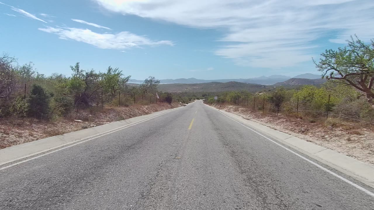 The East Cape Los Cabos road continues through scrubby dry terrain, with a cracked asphalt surface, pale shoulders, low hills in the distance, and no visible people.