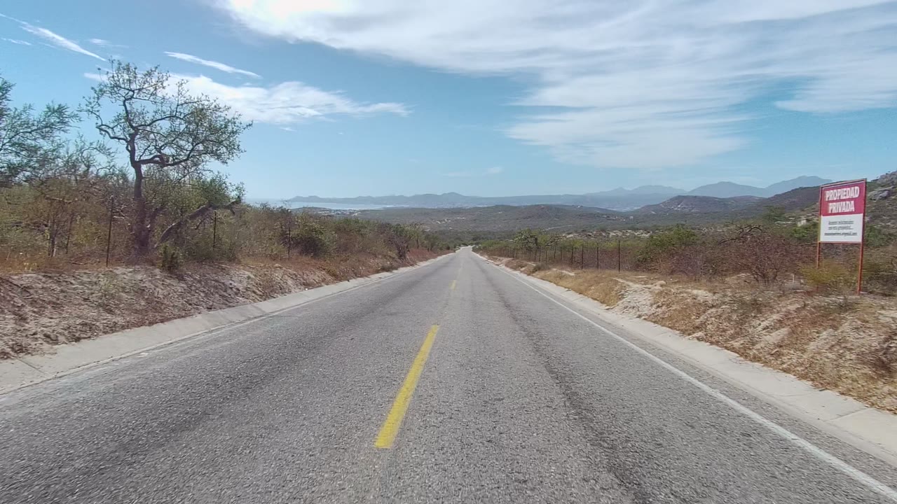 Straight asphalt road in East Cape Los Cabos, Baja California Sur, with dry brush, a red-and-white roadside sign, distant hills, high clouds, and no visible people.