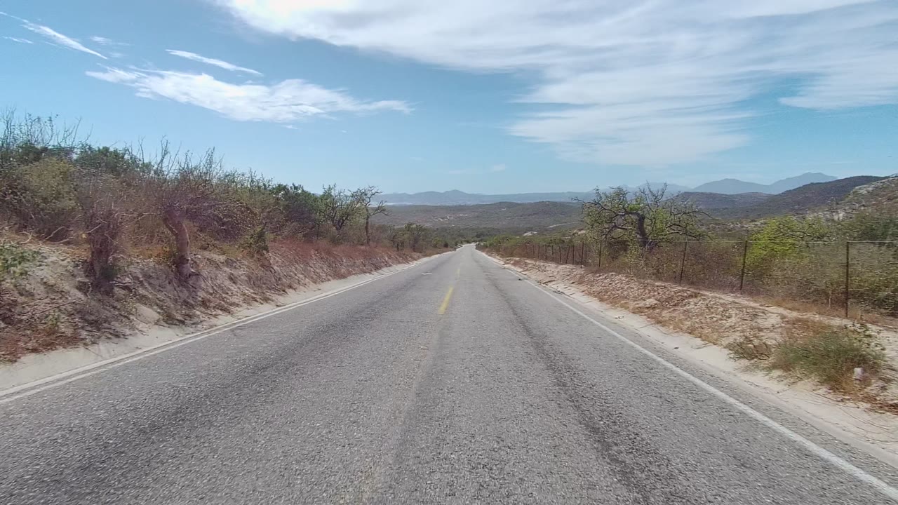 East Cape Los Cabos roadside view with the straight pavement running between sandy shoulders, thorny-looking bushes, wire fencing, low mountains, and no visible people.