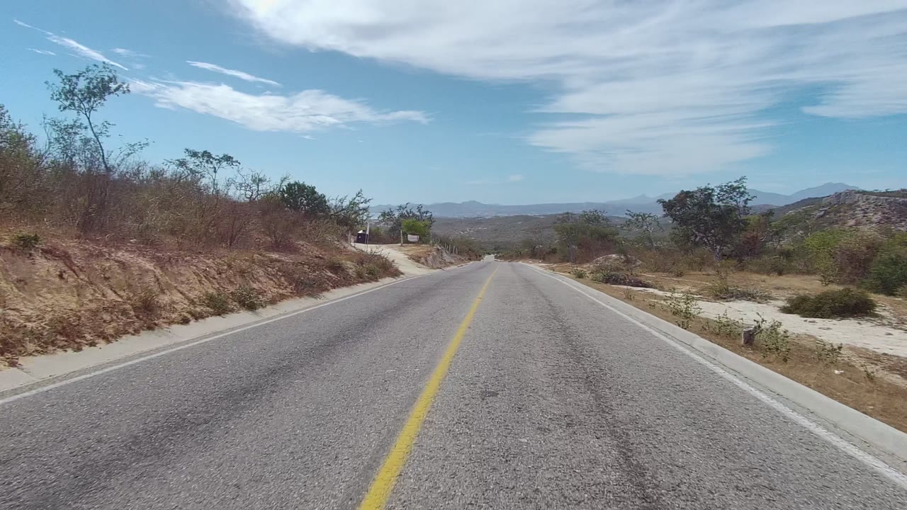 Paved road in East Cape Los Cabos, Baja California Sur, with a yellow center line, sandy shoulders, sparse bushes, distant mountains, and no visible people.