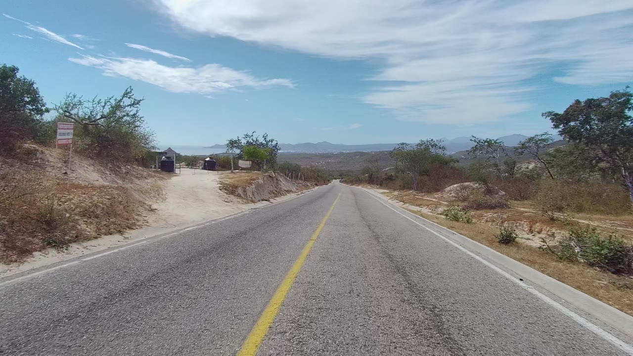 Roadside dirt entrance and sign beside a paved road in arid terrain.