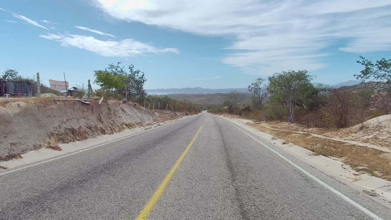 Paved road in East Cape Los Cabos with a raised sandy bank, dry vegetation, small structures set back on the roadside, distant mountains, and no visible people.