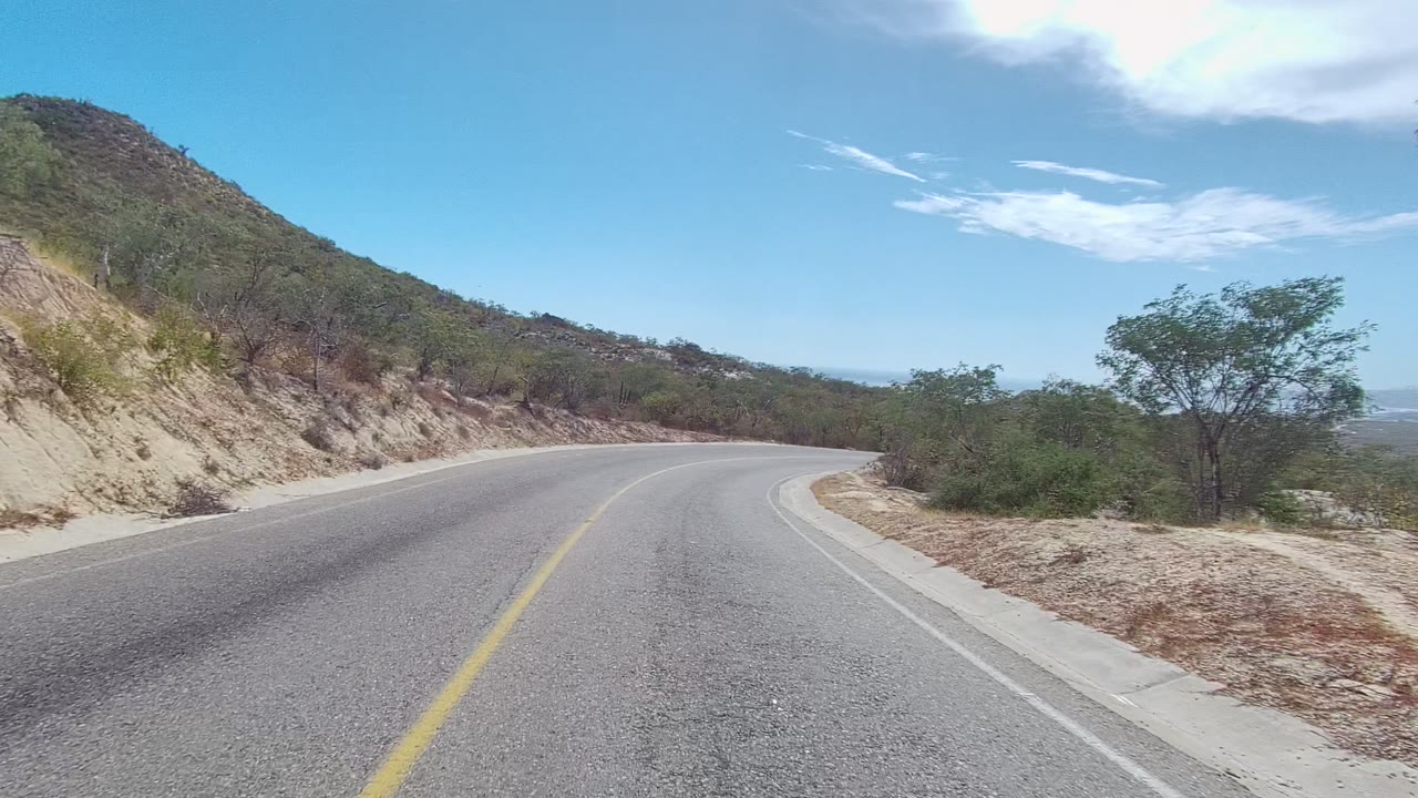 At 0:00, a paved road curves left through an arid hillside in Los Cabos, Baja California Sur, with scrub trees, bright sky, and no visible people.