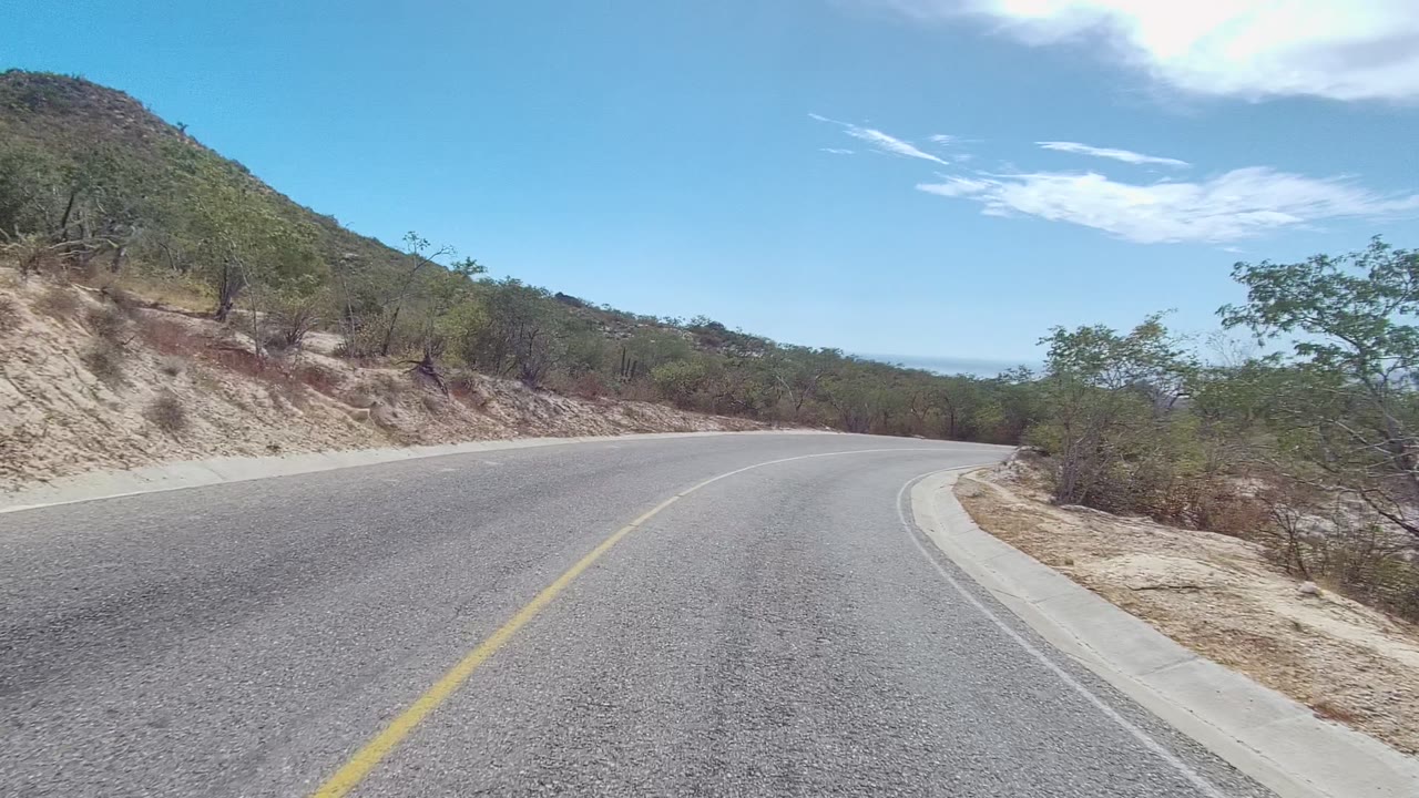 Road bend with yellow line, sandy shoulder, brush, and low hill