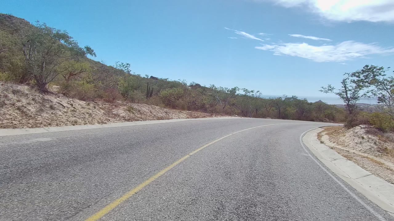 At 0:02, a wider view of the curve shows the asphalt sweeping right through dry brush and low hills in Los Cabos, Baja California Sur, with no visible people.