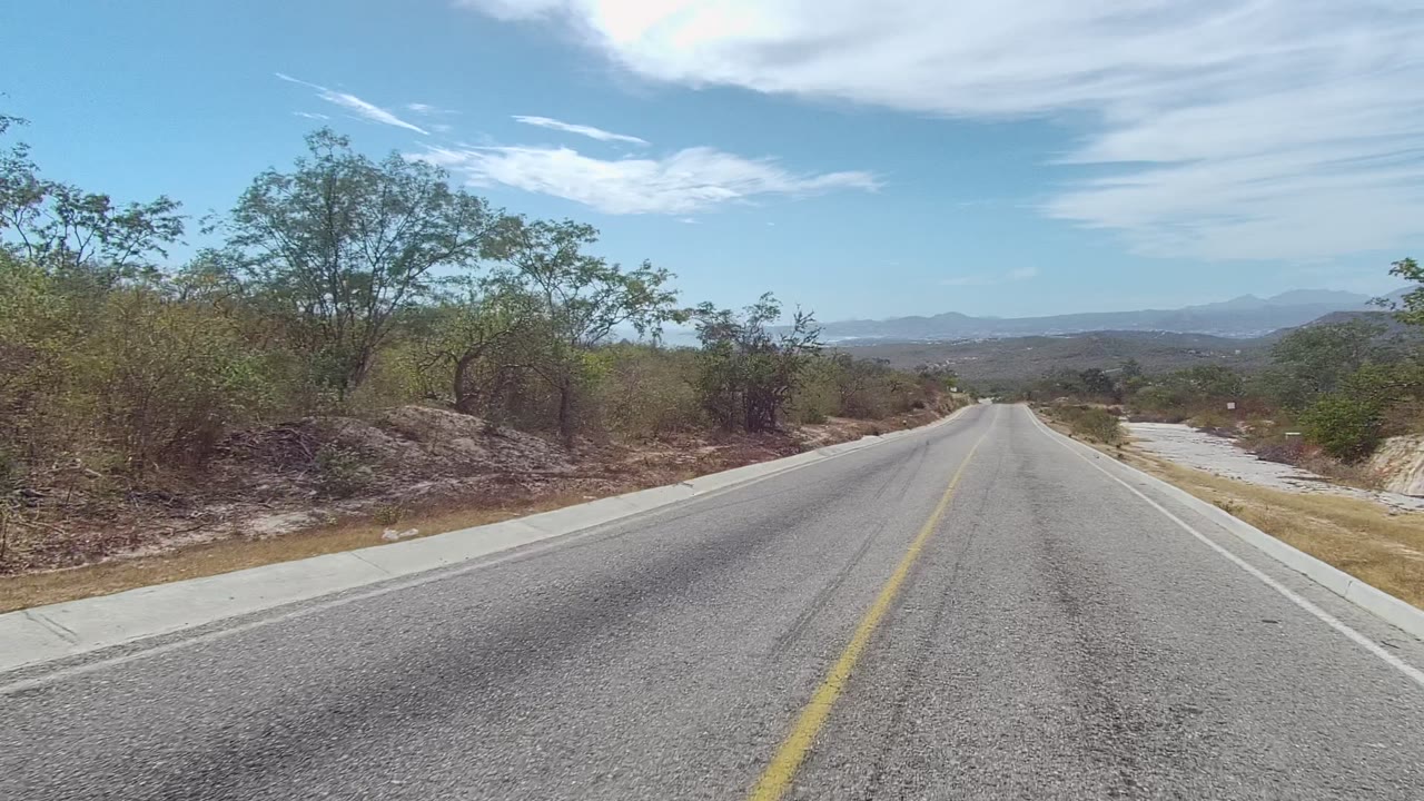 Straight paved road in East Cape Los Cabos, Baja California Sur, with a yellow center line, dry roadside brush, distant low mountains, bright sky, and no visible people.