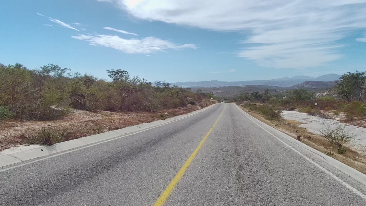 East Cape Los Cabos road scene with the pavement running downhill into dry vegetation, low roadside scrub, distant hills and mountains, scattered white clouds, and no visible people.