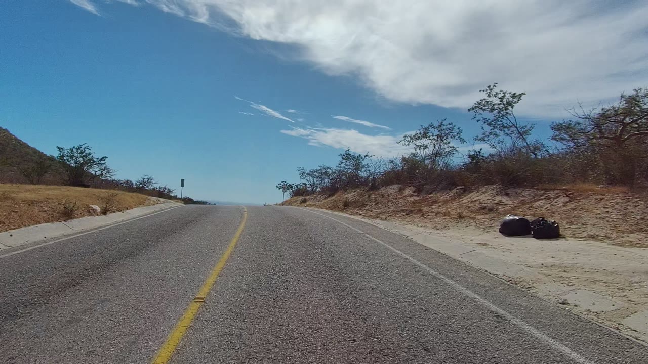 Opening frame of an East Cape Los Cabos paved road with a yellow center line, sandy shoulders, dry hillside brush, large white clouds, and no visible people.
