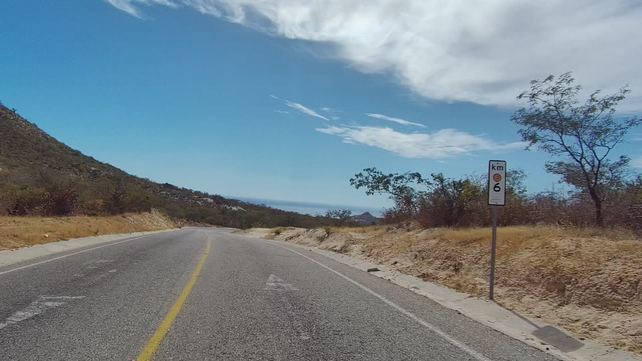 East Cape Los Cabos road curve with a roadside kilometer speed limit sign, arid slope, scattered trees, distant horizon, blue sky, and no visible people.