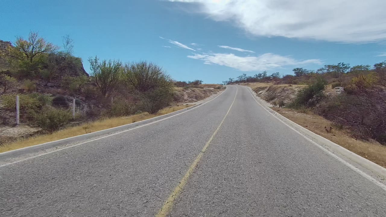 Start of the East Cape Los Cabos road clip showing a two-lane paved road rising gently through dry grass, scattered bushes, low hills, blue sky, and no visible people.