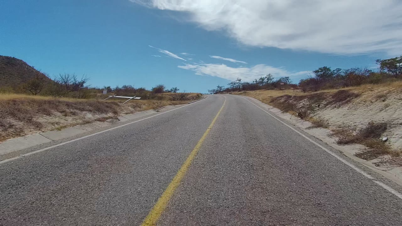 At two seconds, the East Cape Los Cabos roadway fills the foreground while dry embankments, low shrubs, a small roadside post, blue sky, and no visible people remain in view.