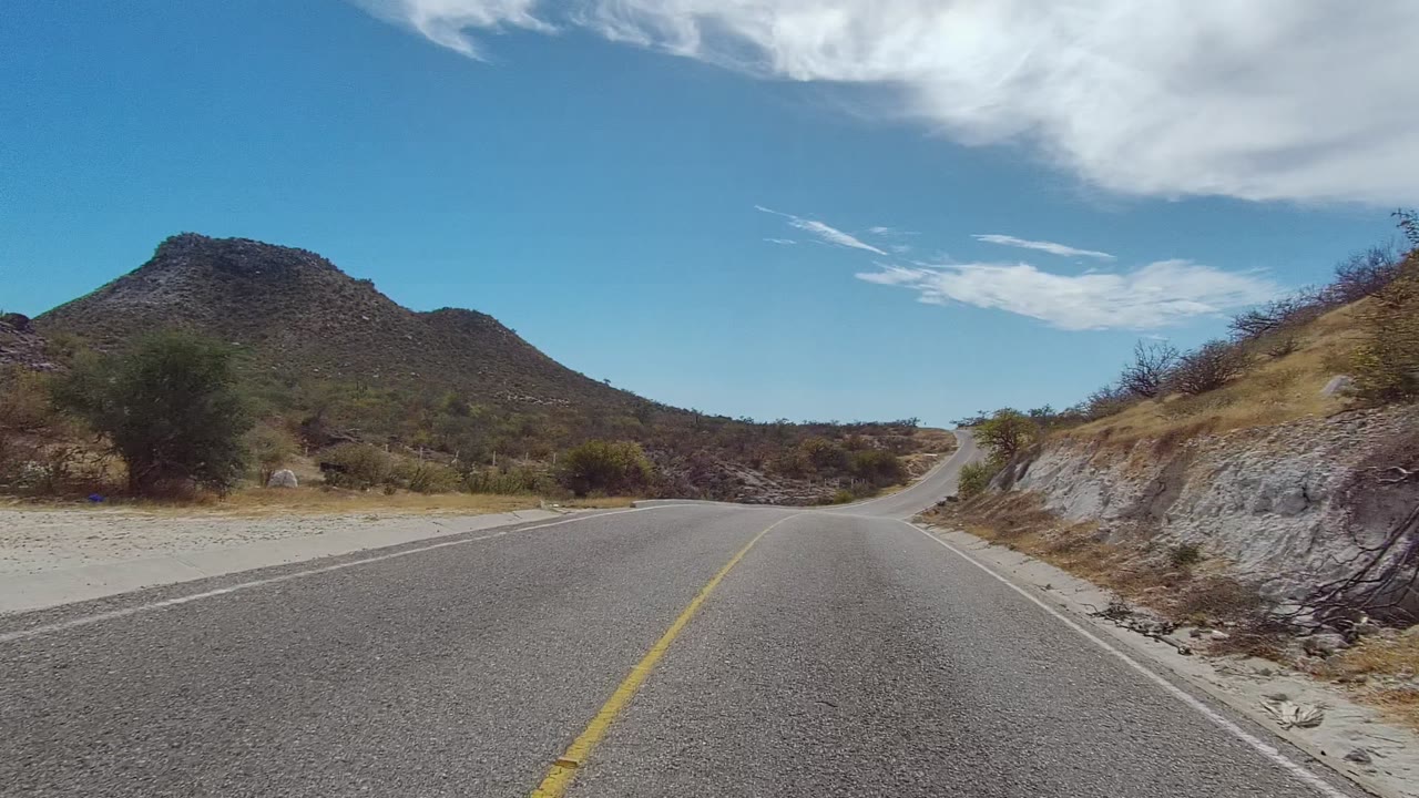 Road-level view in East Cape Los Cabos with a yellow center line leading toward dry brush and a rounded rocky hill, no visible people.