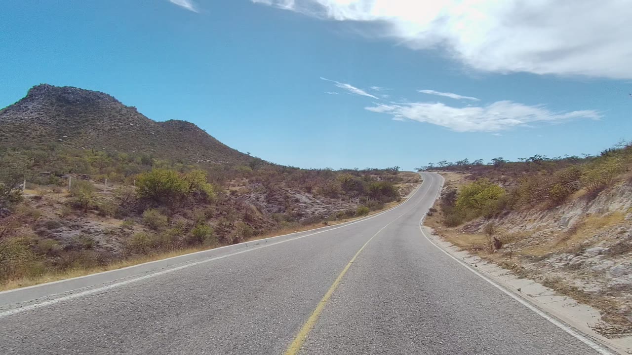 East Cape Los Cabos road scene with the lane rising slightly to the right between rocky slopes, dry brush, and a broad blue sky, no visible people.