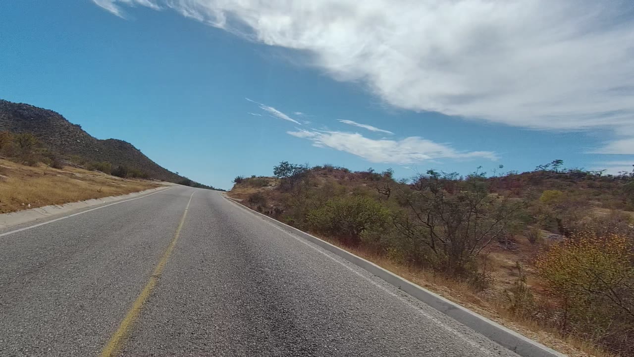 Start of a forward road view in East Cape Los Cabos, with a yellow center line, dry roadside brush, a low mountain at left, blue sky, white cloud cover, and no visible people.