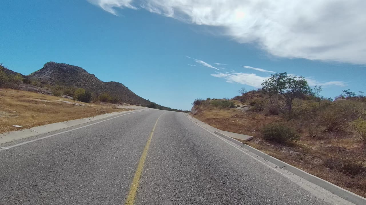 At 0:02, the East Cape Los Cabos road leads past sparse desert vegetation, with the yellow line bending slightly, a mountain ahead-left, broad white clouds overhead, and no visible people.