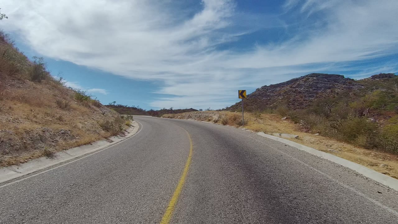 A paved road curves left through dry hills in East Cape Los Cabos, Baja California Sur, with a yellow curve sign ahead, blue sky, white clouds, and no visible people.